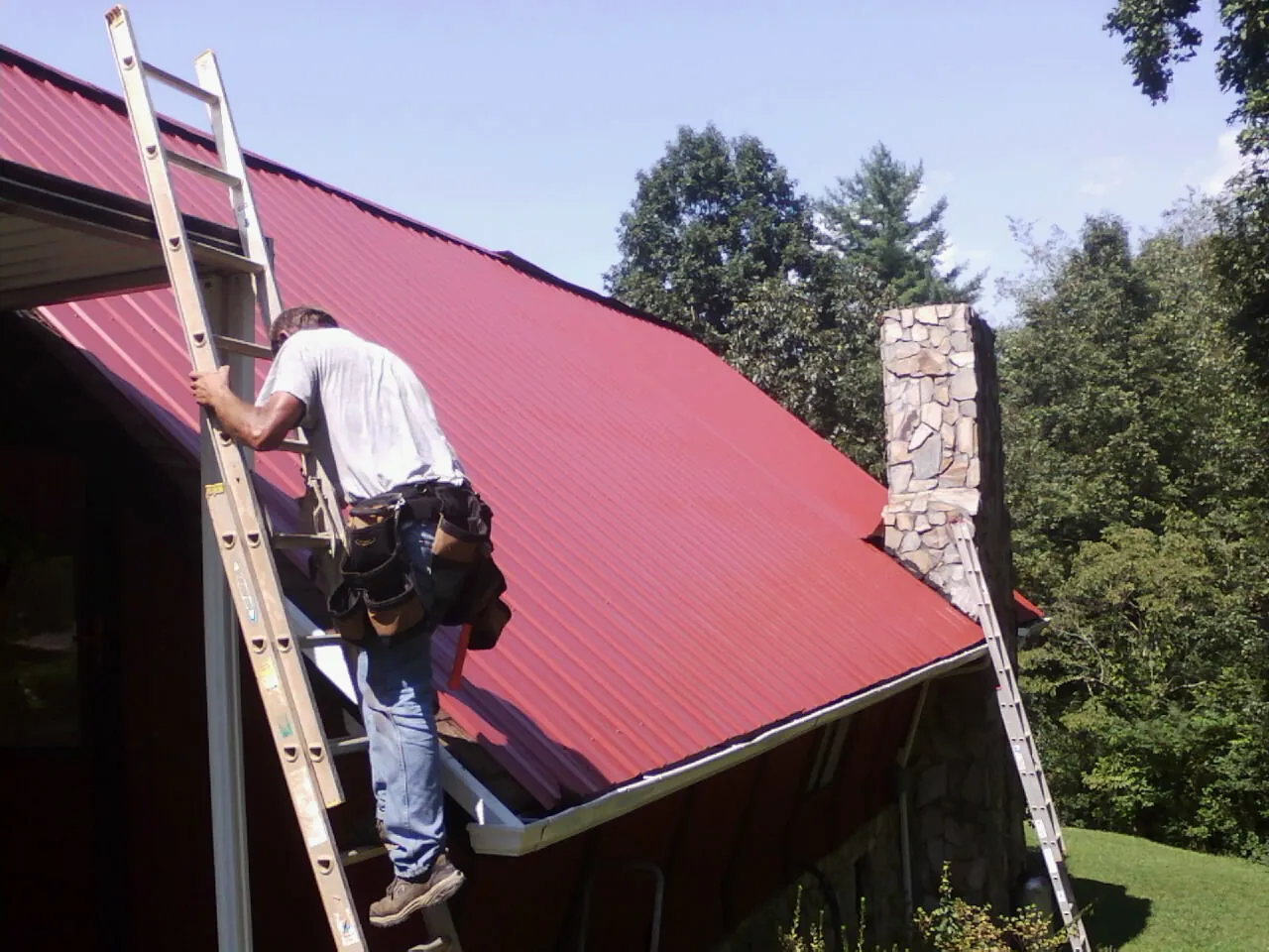 Roofer on a ladder beside a red metal roof and stone chimney — Soffit & Fascia Repair by Roofing Brothers in Florissant